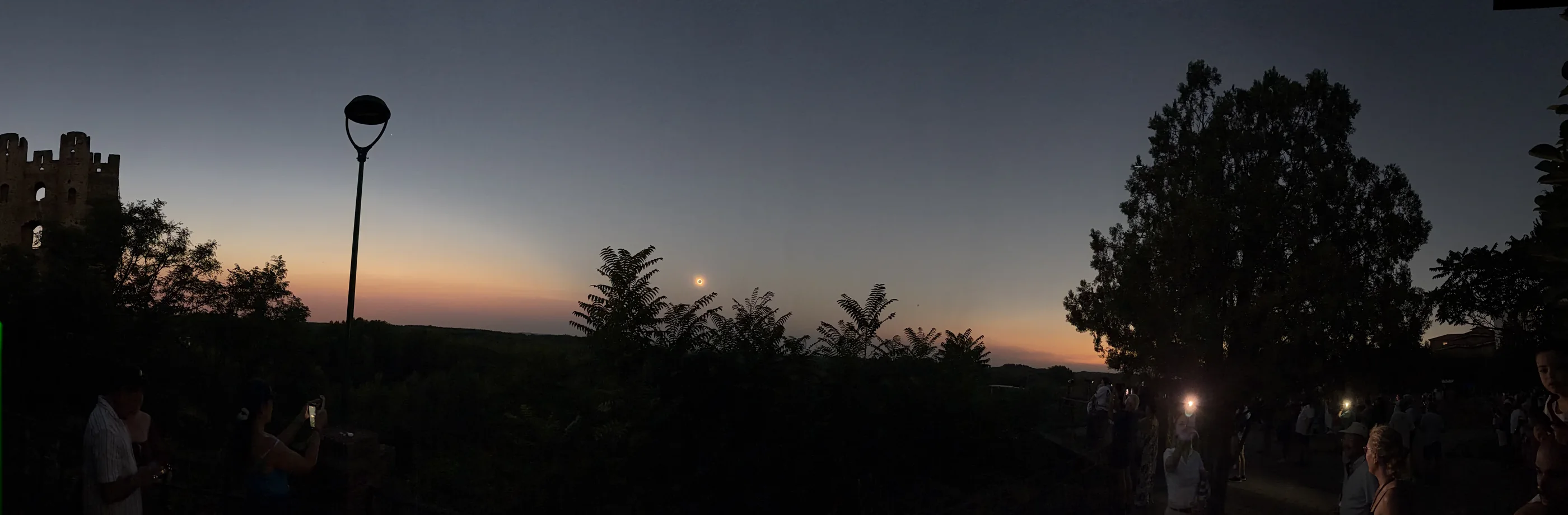 Wide panorama during totality showing the eclipsed Sun above a dark horizon with orange illuminated atmosphere on both sides